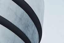 Curved architectural structure featuring overlapping layers with a smooth concrete surface, captured against a light blue sky.