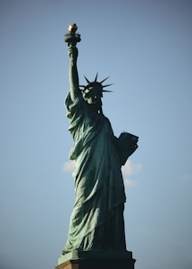 A towering statue of a robed female figure holding a torch in her raised right hand and a book in her left. The figure is adorned with a spiked crown and stands on a pedestal against a clear blue sky.