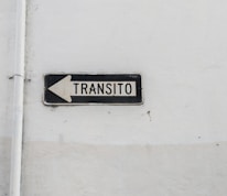A weathered traffic sign with black text 'TRANSITO' and a left-pointing arrow is mounted against a plain, worn white wall. The sign is rectangular with a black border and white background.