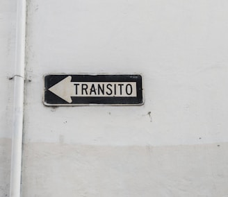 A weathered traffic sign with black text 'TRANSITO' and a left-pointing arrow is mounted against a plain, worn white wall. The sign is rectangular with a black border and white background.