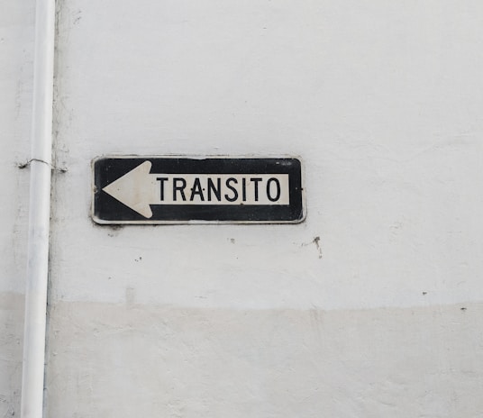 A weathered traffic sign with black text 'TRANSITO' and a left-pointing arrow is mounted against a plain, worn white wall. The sign is rectangular with a black border and white background.