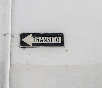A weathered traffic sign with black text 'TRANSITO' and a left-pointing arrow is mounted against a plain, worn white wall. The sign is rectangular with a black border and white background.
