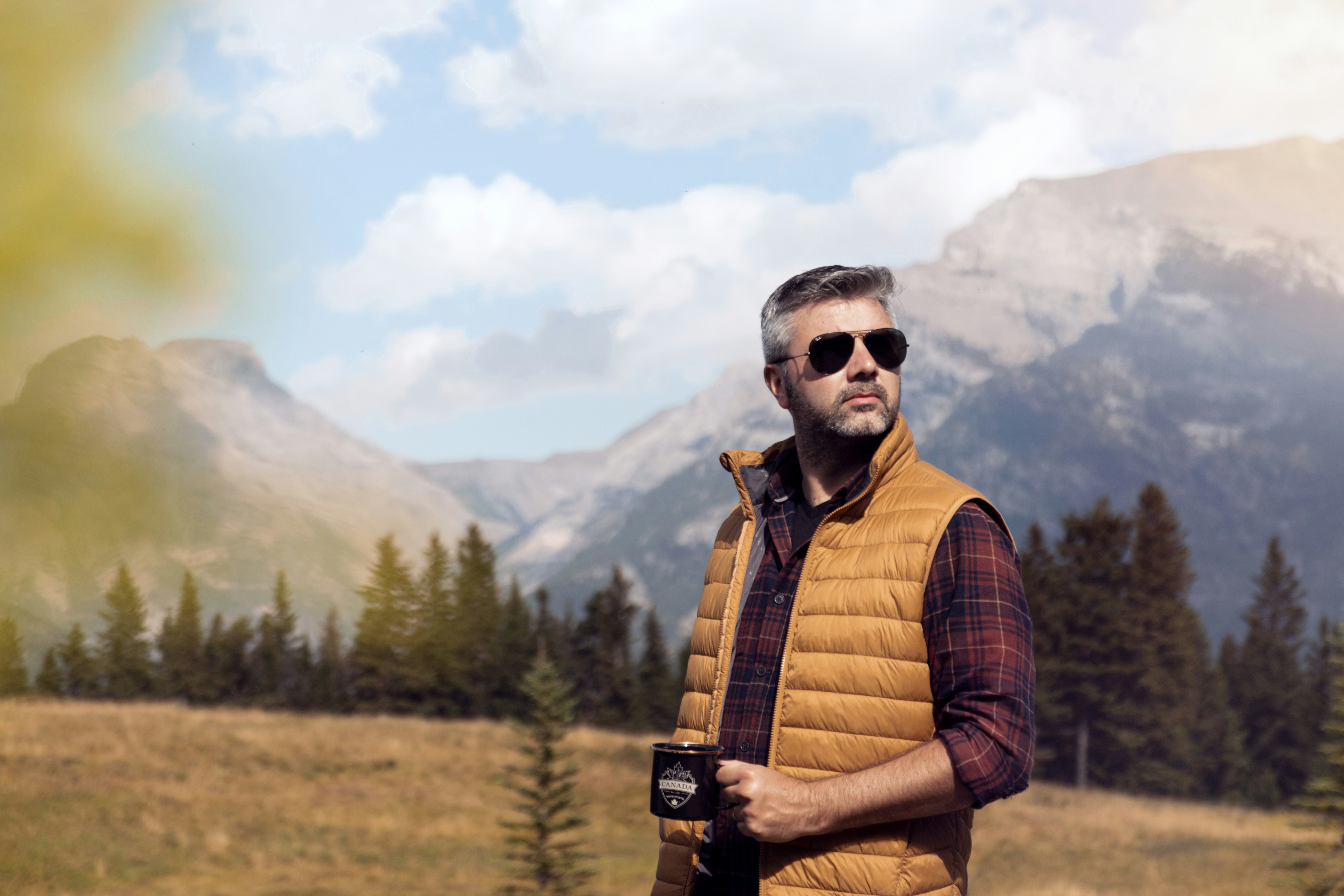 Gray-haired man in sunglasses, a tan quilted puffer vest and a plaid flannel shirt holding a mug in a mountain meadow, snow on the peaks behind him