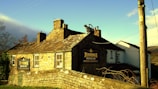 A rustic stone inn with the name 'Jennings Bridge Inn' displayed on signs mounted on the building's exterior. The inn is constructed with a traditional design featuring chimney stacks and a sloped roof. A decorative gargoyle is positioned on the roof. The building is surrounded by a stone wall, and a utility pole is visible on the right. The surrounding area includes bare trees and there is a clear blue sky with some clouds.