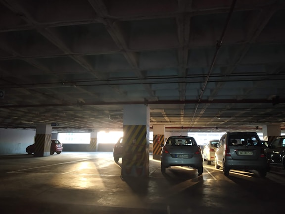 A dimly-lit parking garage with several cars parked within marked spaces. The structure is characterized by concrete columns and beams, and sunlight filters in from an opening in the background, casting elongated shadows on the floor.