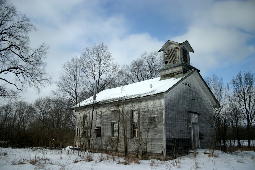 gray wooden house near snowy field surrounded with bare trees under white and blue sky during daytime