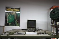 A retro film editing machine with various knobs and a small screen is positioned centrally. To the left, a poster of the classic film 'Metropolis' features prominently on the wall, depicting a stylized green figure with a futuristic helmet. On the right, there is a large vintage spotlight, and a bookshelf is partially visible.