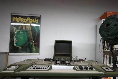 A retro film editing machine with various knobs and a small screen is positioned centrally. To the left, a poster of the classic film 'Metropolis' features prominently on the wall, depicting a stylized green figure with a futuristic helmet. On the right, there is a large vintage spotlight, and a bookshelf is partially visible.