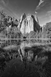 A black and white photograph depicts a rugged mountain range with steep cliffs. The mountains are mirrored in the calm water below, surrounded by dense forest and tall trees. The sky above is mostly clear with a few wispy clouds.