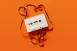 A vintage white cassette tape is placed on an orange background. The tape is partially unraveled, with the ribbon forming loose, swirling patterns around the cassette.