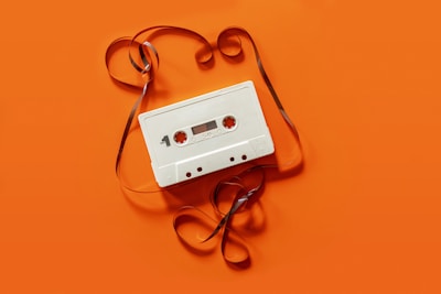 A vintage white cassette tape is placed on an orange background. The tape is partially unraveled, with the ribbon forming loose, swirling patterns around the cassette.