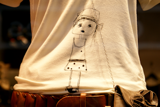 A white shirt with a simple black line drawing of a stick figure in a dress. The figure has simple features, including eyes and a hat. A brown belt is worn around the waist, and a purse with a chain strap hangs from the shoulder.