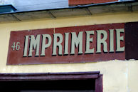 A weathered sign with the word 'IMPRIMERIE' prominently displayed in bold, serif letters on a red background. The number 46 is positioned to the left of the text. The sign is mounted on a light yellow wall, which appears to be part of an older building.