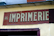 A weathered sign with the word 'IMPRIMERIE' prominently displayed in bold, serif letters on a red background. The number 46 is positioned to the left of the text. The sign is mounted on a light yellow wall, which appears to be part of an older building.