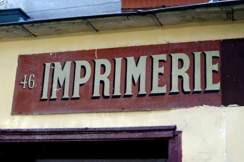 A weathered sign with the word 'IMPRIMERIE' prominently displayed in bold, serif letters on a red background. The number 46 is positioned to the left of the text. The sign is mounted on a light yellow wall, which appears to be part of an older building.
