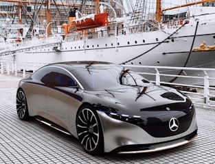 A sleek, modern luxury car with a shiny black and silver finish is parked on a tiled surface. The background features a large, white ship with visible masts and rigging, suggesting a marina or port setting. The contrast between the contemporary design of the vehicle and the classic ship creates a dynamic visual impact.