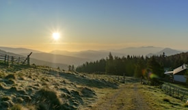 A tranquil sunrise over a mountainous landscape with a gentle sun filtering through the misty sky. A grassy path leads towards a dense forest of evergreen trees. A rustic wooden fence and a small cabin are visible, adding to the serene countryside setting.