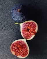 A whole dark purple fig sits beside a ripe, halved fig. The interior of the cut fig reveals a complex texture of red and pink flesh dotted with small seeds. The figs rest on a dark textured surface, highlighting their vibrant colors.