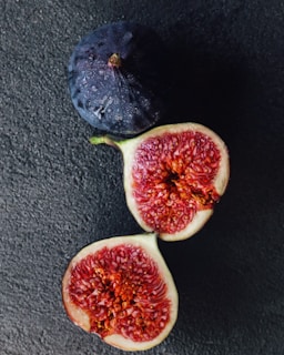 A whole dark purple fig sits beside a ripe, halved fig. The interior of the cut fig reveals a complex texture of red and pink flesh dotted with small seeds. The figs rest on a dark textured surface, highlighting their vibrant colors.