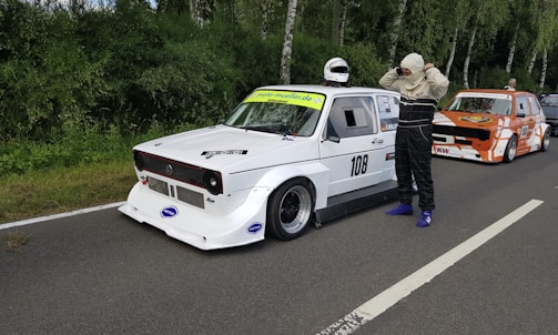 A white race car with the number 108 is parked on a road with a driver standing nearby. The driver, dressed in racing gear, is adjusting their headgear. Another car, painted in orange and white, is positioned behind it. Both cars are modified for racing, with aerodynamic features. The scene is set against a backdrop of green trees along the roadside.