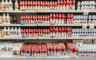 A supermarket shelf displays a variety of yogurt bottles arranged in neat rows. The bottles feature vibrant packaging, predominantly in red and white, with images of fruits such as berries and peaches. Price tags are visible on the shelves, indicating the cost of each product.