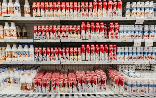 A supermarket shelf displays a variety of yogurt bottles arranged in neat rows. The bottles feature vibrant packaging, predominantly in red and white, with images of fruits such as berries and peaches. Price tags are visible on the shelves, indicating the cost of each product.