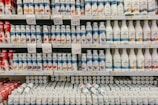 Rows of various flavored yogurt bottles are neatly arranged on supermarket shelves, featuring several brands and flavors. Some bottles are white while others are colorful with images of fruits like strawberries, blueberries, and other ingredients. Price tags are displayed prominently above the products.