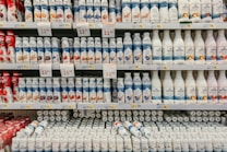 Rows of various flavored yogurt bottles are neatly arranged on supermarket shelves, featuring several brands and flavors. Some bottles are white while others are colorful with images of fruits like strawberries, blueberries, and other ingredients. Price tags are displayed prominently above the products.