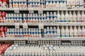 Rows of various flavored yogurt bottles are neatly arranged on supermarket shelves, featuring several brands and flavors. Some bottles are white while others are colorful with images of fruits like strawberries, blueberries, and other ingredients. Price tags are displayed prominently above the products.
