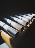 A series of cylindrical metal pipes arranged horizontally, casting shadows on a wooden surface. The lighting creates striking contrasts between the bright highlights on the pipes and the dark shadows.