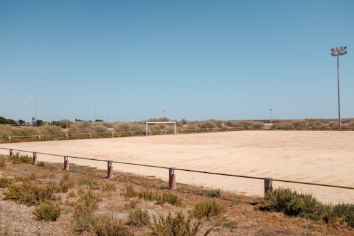 An empty, sandy sports field surrounded by tall grass and a few floodlights. A single goalpost stands at one end, while sparse vegetation lines the perimeter. The sky is clear blue, suggesting a bright, sunny day.