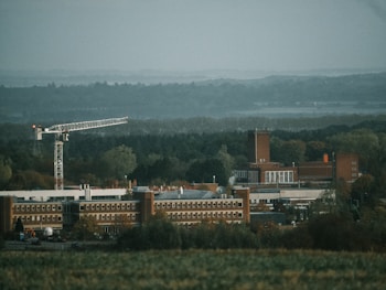 A large industrial building complex surrounded by dense forest. A tall construction crane is positioned prominently to the left, indicating ongoing development. The buildings have a brick exterior with multiple windows and a tall chimney-like structure. The landscape features a green, wooded area under an overcast sky.