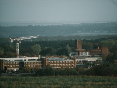 A large industrial building complex surrounded by dense forest. A tall construction crane is positioned prominently to the left, indicating ongoing development. The buildings have a brick exterior with multiple windows and a tall chimney-like structure. The landscape features a green, wooded area under an overcast sky.