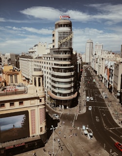 An urban scene featuring a prominent art deco building with the word 'Schweppes' displayed on its curved facade. The architecture blends into a bustling cityscape with a wide avenue extending towards the horizon, flanked by additional buildings with classic designs. Pedestrians and vehicles populate the street, while a billboard advertising 'Cines Callao' adds to the metropolitan atmosphere. The sky is partly cloudy, enhancing the warm tones of the city.