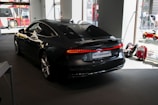 A sleek black Audi A7 Sportback is positioned in a well-lit showroom. The vehicle's streamlined design is highlighted under the indoor lighting, emphasizing its glossy finish. Outside the large windows, a red double-decker bus is partially visible, along with a toy car and a red suitcase placed next to the car, suggesting a showroom or exhibition setting.
