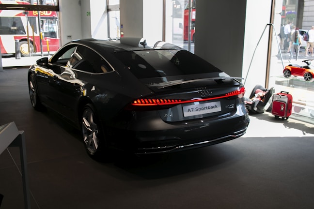 A sleek black Audi A7 Sportback is positioned in a well-lit showroom. The vehicle's streamlined design is highlighted under the indoor lighting, emphasizing its glossy finish. Outside the large windows, a red double-decker bus is partially visible, along with a toy car and a red suitcase placed next to the car, suggesting a showroom or exhibition setting.