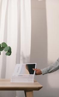 A stack of magazines titled 'Genesis' is neatly placed on a wooden table. A hand, appearing from the right, is picking up the top magazine. Green leaves from a plant extend from the left, contrasting against a white curtain in the background.