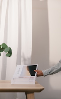 A stack of magazines titled 'Genesis' is neatly placed on a wooden table. A hand, appearing from the right, is picking up the top magazine. Green leaves from a plant extend from the left, contrasting against a white curtain in the background.