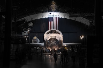 A space shuttle is prominently displayed in a museum-like setting. The American flag hangs above it, and various aerospace exhibits surround the area. The environment is dimly lit, creating a dramatic focus on the shuttle. Several visitors are walking around, observing the exhibit.