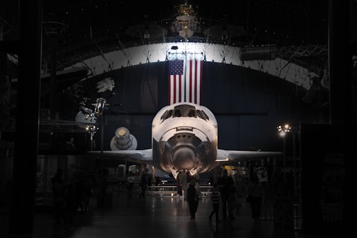 A space shuttle is prominently displayed in a museum-like setting. The American flag hangs above it, and various aerospace exhibits surround the area. The environment is dimly lit, creating a dramatic focus on the shuttle. Several visitors are walking around, observing the exhibit.