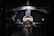 A space shuttle is prominently displayed in a museum-like setting. The American flag hangs above it, and various aerospace exhibits surround the area. The environment is dimly lit, creating a dramatic focus on the shuttle. Several visitors are walking around, observing the exhibit.