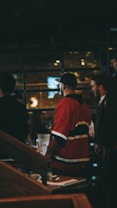 A group of people are indoors, with one person prominently wearing a red sports jersey and a cap. The setting appears to be either a bar or a restaurant with dim lighting and visible wooden furniture. There is a blurred background with lights and possibly windows.