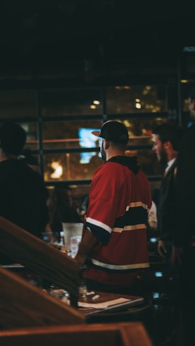 A group of people are indoors, with one person prominently wearing a red sports jersey and a cap. The setting appears to be either a bar or a restaurant with dim lighting and visible wooden furniture. There is a blurred background with lights and possibly windows.