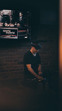 A person wearing a hat is focused on operating DJ equipment in a dimly lit room with a brick wall. Above this setup, a television screen displays a broadcast featuring three individuals discussing sports.