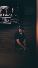 A person wearing a hat is focused on operating DJ equipment in a dimly lit room with a brick wall. Above this setup, a television screen displays a broadcast featuring three individuals discussing sports.