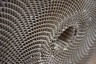 A close-up view of a corrugated cardboard material, showcasing its intricate honeycomb-like pattern. The image highlights the symmetrical, repetitive structure of the corrugated layers, creating a sense of depth and texture.