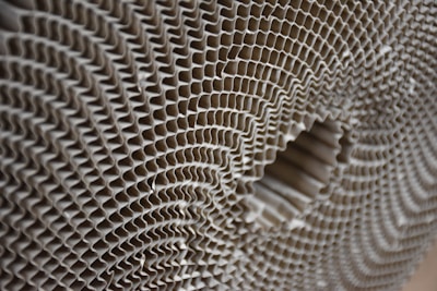 A close-up view of a corrugated cardboard material, showcasing its intricate honeycomb-like pattern. The image highlights the symmetrical, repetitive structure of the corrugated layers, creating a sense of depth and texture.