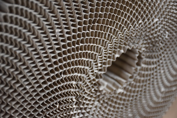A close-up view of a corrugated cardboard material, showcasing its intricate honeycomb-like pattern. The image highlights the symmetrical, repetitive structure of the corrugated layers, creating a sense of depth and texture.