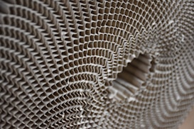 A close-up view of a corrugated cardboard material, showcasing its intricate honeycomb-like pattern. The image highlights the symmetrical, repetitive structure of the corrugated layers, creating a sense of depth and texture.