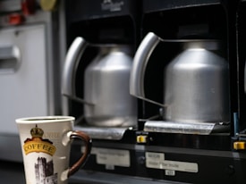 A close-up view of two stainless steel coffee carafes placed in a machine, with a ceramic coffee mug featuring a decorative design and the word 'COFFEE' written on it in the foreground.